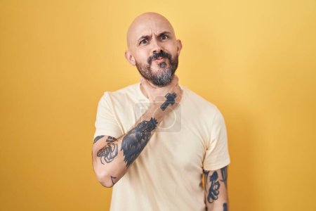 Téléchargez les photos : Homme hispanique avec des tatouages debout sur fond jaune couper la gorge avec la main comme couteau, menacer d'agression avec une violence furieuse - en image libre de droit