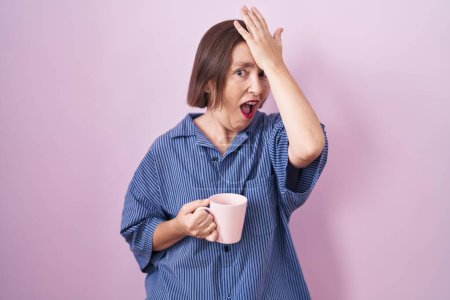 Foto de Mujer hispana de mediana edad bebiendo una taza de café sorprendida con la mano en la cabeza por error, recuerde el error. olvidado, mal concepto de memoria. - Imagen libre de derechos