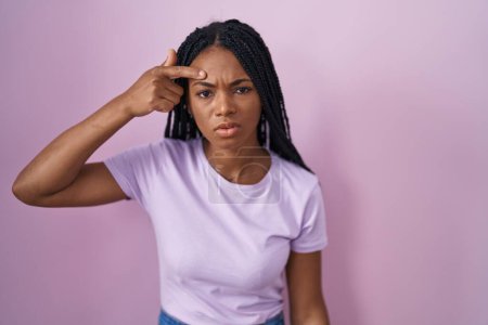 Foto de Mujer afroamericana con trenzas de pie sobre fondo rosa apuntando infeliz a la espinilla en la frente, fea infección de la cabeza negra. acné y problemas de piel - Imagen libre de derechos