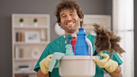 Téléchargez les photos : Jeune homme hispanique nettoyeur professionnel tenant des produits propres souriant à la maison - en image libre de droit