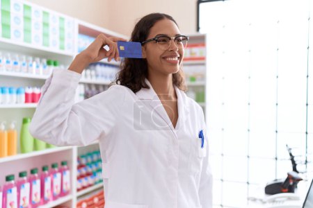 Téléchargez les photos : Jeune pharmacienne afro-américaine souriante confiante tenant sa carte de crédit à la pharmacie - en image libre de droit