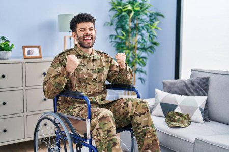 Foto de Hombre árabe vistiendo camuflaje uniforme del ejército sentado en silla de ruedas muy feliz y emocionado haciendo gesto ganador con los brazos levantados, sonriendo y gritando por el éxito. concepto de celebración. - Imagen libre de derechos