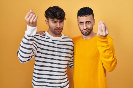 Foto de Joven pareja gay hispana de pie sobre fondo amarillo haciendo gesto italiano con la mano y los dedos expresión confiada - Imagen libre de derechos