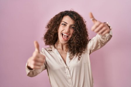 Foto de Mujer hispana con el pelo rizado de pie sobre fondo rosa aprobando hacer gesto positivo con la mano, pulgares hacia arriba sonriendo y feliz por el éxito. gesto ganador. - Imagen libre de derechos