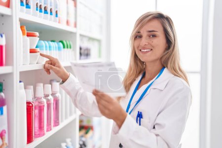 Téléchargez les photos : Jeune femme blonde pharmacien lecture prescription à la pharmacie - en image libre de droit