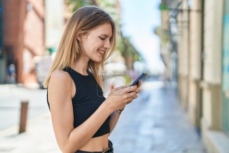 Photo for Young blonde woman using smartphone smiling at street - Royalty Free Image