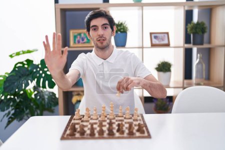 Téléchargez les photos : Jeune homme hispanique jouant aux échecs assis sur la table avec la main ouverte faisant signe d'arrêt avec expression sérieuse et confiante, geste de défense - en image libre de droit