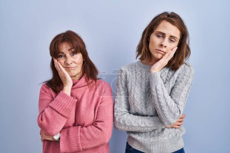 Mother and daughter standing over blue background thinking looking tired and bored with depression problems with crossed arms. 