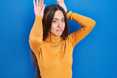 Photo for Young brunette woman standing over blue background doing bunny ears gesture with hands palms looking cynical and skeptical. easter rabbit concept. - Royalty Free Image