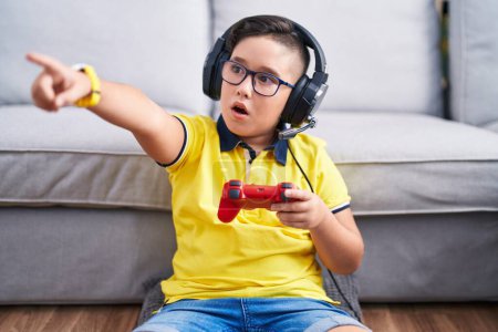 Foto de Joven niño hispano jugando videojuego sosteniendo controlador usando auriculares apuntando con el dedo sorprendido por delante, boca abierta expresión asombrada, algo en el frente - Imagen libre de derechos