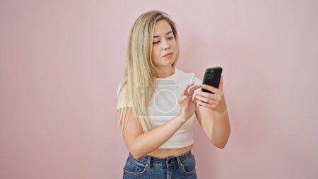 Foto de Mujer rubia joven usando smartphone con expresión seria sobre fondo rosa aislado - Imagen libre de derechos