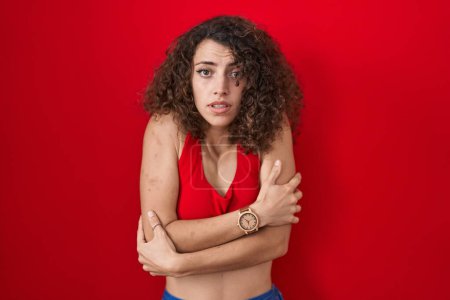 Photo for Hispanic woman with curly hair standing over red background shaking and freezing for winter cold with sad and shock expression on face - Royalty Free Image