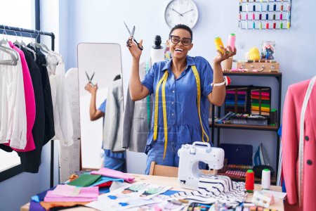 Téléchargez les photos : Femme afro-américaine tailleur souriant confiant tenant des ciseaux et du fil à tailleur magasin - en image libre de droit