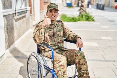 Téléchargez les photos : Jeune homme arabe portant un uniforme de camouflage de l'armée assis sur un fauteuil roulant souriant heureux et positif, pouce levé faire excellent et signe d'approbation - en image libre de droit