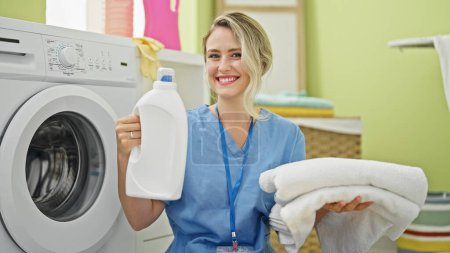 Téléchargez les photos : Jeune femme blonde nettoyant professionnel tenant bouteille de détergent et serviettes pliées à la buanderie - en image libre de droit