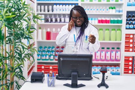 Photo for African american woman pharmacist holding pills bottle talking on smartphone at pharmacy - Royalty Free Image