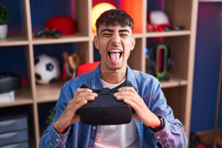 Foto de Joven hombre hispano con gafas de realidad virtual sacando la lengua feliz con expresión divertida. - Imagen libre de derechos