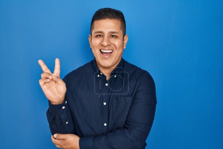 Téléchargez les photos : Jeune homme hispanique debout sur fond bleu souriant avec un sourire heureux clin d'oeil à la caméra faisant signe de victoire. numéro deux. - en image libre de droit