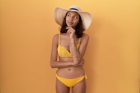 Foto de Mujer hispana joven con bikini y sombrero de verano mirando con confianza a la cámara sonriendo con los brazos cruzados y la mano levantada en la barbilla. pensar positivamente. - Imagen libre de derechos