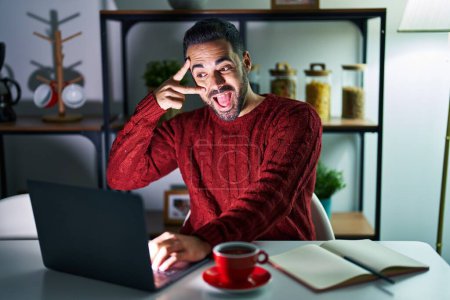 Téléchargez les photos : Jeune homme hispanique avec barbe utilisant ordinateur portable la nuit à la maison faisant symbole de paix avec les doigts sur le visage, souriant gai montrant la victoire - en image libre de droit