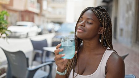 Téléchargez les photos : Femme afro-américaine tenant un verre d'eau assis sur la table à la terrasse du café - en image libre de droit