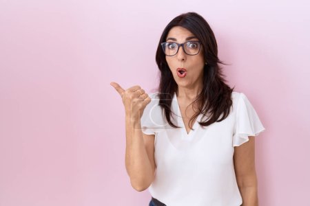 Foto de Mujer hispana de mediana edad vistiendo camiseta blanca casual y gafas sorprendidas apuntando con el dedo de la mano hacia un lado, boca abierta expresión asombrada. - Imagen libre de derechos