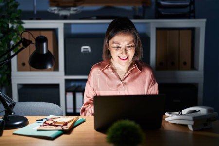 Téléchargez les photos : Moyen Age femme hispanique travaillant à l'aide d'un ordinateur portable tard dans la nuit regardant positif et heureux debout et souriant avec un sourire confiant montrant les dents - en image libre de droit