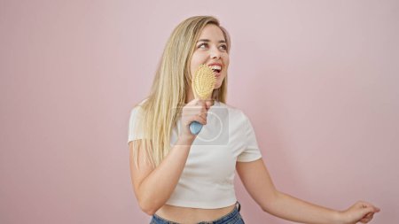 Téléchargez les photos : Jeune femme blonde chantant chanson en utilisant peigne comme un microphone sur fond rose isolé - en image libre de droit