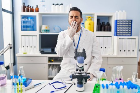 Foto de Un joven hispano con barba trabajando en un laboratorio científico aburrido bostezando cansado cubriendo la boca con la mano. inquietud y somnolencia. - Imagen libre de derechos