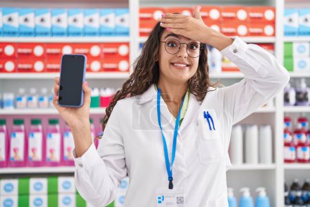 Téléchargez les photos : Jeune femme hispanique travaillant à la pharmacie pharmacie montrant écran smartphone langue collante heureux avec expression drôle. - en image libre de droit