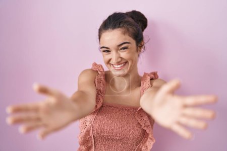 Foto de Joven adolescente de pie sobre fondo rosa mirando a la cámara sonriendo con los brazos abiertos para el abrazo. expresión alegre abrazando la felicidad. - Imagen libre de derechos