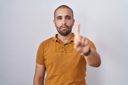 Foto de Hombre hispano con barba de pie sobre fondo blanco señalando con el dedo hacia arriba y expresión enojada, sin mostrar ningún gesto - Imagen libre de derechos