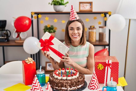 Foto de Mujer hispana celebrando cumpleaños con pastel sosteniendo regalo sonriendo con una sonrisa feliz y fresca en la cara. mostrando dientes. - Imagen libre de derechos