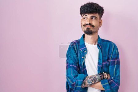 Foto de Joven hombre hispano con barba de pie sobre fondo rosa sonriendo mirando hacia un lado y mirando hacia otro pensando. - Imagen libre de derechos