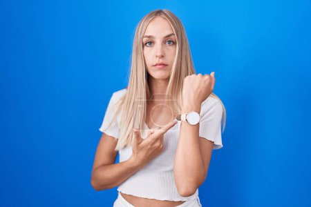 Téléchargez les photos : Young caucasian woman standing over blue background in hurry pointing to watch time, impatience, looking at the camera with relaxed expression - en image libre de droit