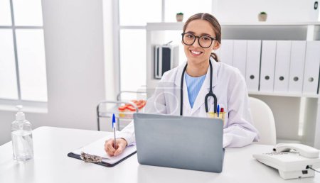 Photo for Young beautiful hispanic woman doctor using laptop writing medical report at clinic - Royalty Free Image