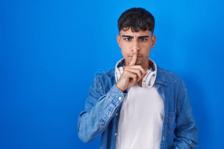 Téléchargez les photos : Jeune homme hispanique debout sur fond bleu demandant à se taire avec le doigt sur les lèvres. silence et concept secret. - en image libre de droit