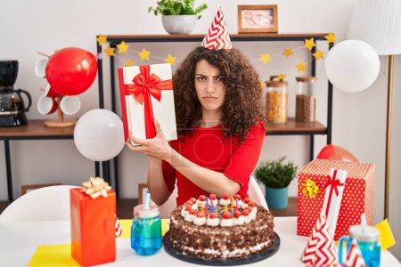 Téléchargez les photos : Hispanic woman with curly hair celebrating birthday with cake and present skeptic and nervous, frowning upset because of problem. negative person. - en image libre de droit