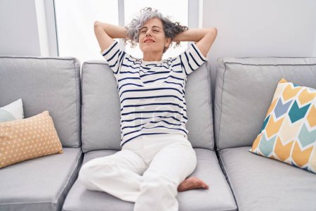 Téléchargez les photos : Femme aux cheveux gris d'âge moyen détendue avec les mains sur la tête assise sur le canapé à la maison - en image libre de droit