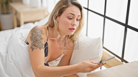 Foto de Mujer rubia joven acostada en la cama mirando teléfono inteligente sorprendido en el dormitorio - Imagen libre de derechos