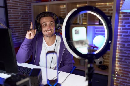 Téléchargez les photos : Homme hispanique avec barbe jouant à des jeux vidéo enregistrement avec smartphone souriant avec une idée ou une question pointant du doigt avec un visage heureux, numéro un - en image libre de droit