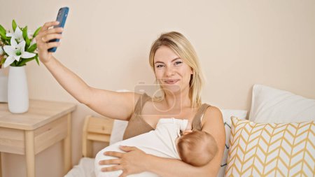 Foto de Madre e hija sentadas en la cama amamantando al bebé hacen selfie por teléfono inteligente en el dormitorio - Imagen libre de derechos