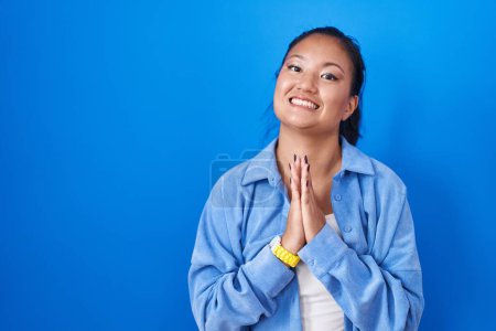 Téléchargez les photos : Asiatique jeune femme debout sur fond bleu priant avec les mains ensemble demandant pardon souriant confiant. - en image libre de droit