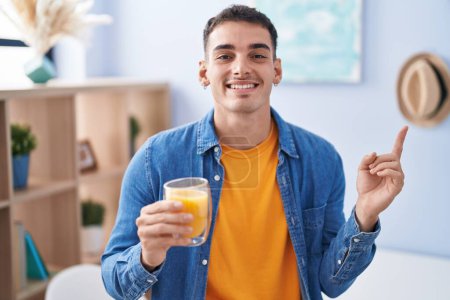 Téléchargez les photos : Bel homme hispanique boire verre de jus d'orange souriant heureux pointant avec la main et le doigt sur le côté - en image libre de droit