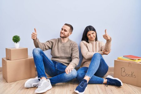 Téléchargez les photos : Jeune couple déménageant dans une nouvelle maison à l'air fier, souriant faire les pouces vers le haut geste sur le côté - en image libre de droit