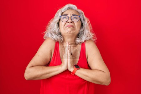 Téléchargez les photos : Femme d'âge moyen avec les cheveux gris debout sur fond rouge mendier et prier avec les mains ainsi que l'expression de l'espoir sur le visage très émotionnel et inquiet. mendier. - en image libre de droit