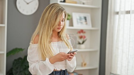 Téléchargez les photos : Young blonde woman using smartphone standing at home - en image libre de droit