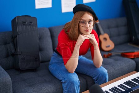 Téléchargez les photos : Jeune femme rousse artiste assise sur un canapé avec une expression sérieuse au studio de musique - en image libre de droit