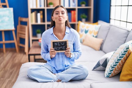 Photo for Young hispanic woman holding blackboard with new home text sitting on the sofa looking at the camera blowing a kiss being lovely and sexy. love expression. - Royalty Free Image