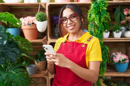 Téléchargez les photos : Jeune belle femme arabe fleuriste souriant confiant en utilisant un smartphone à la boutique de fleurs - en image libre de droit